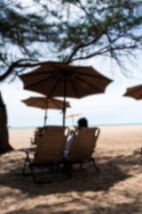Blurred background of man sitting chair along the shoreline with sun umbrella, evoking a sense of relaxation and escape