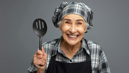 Elderly woman in checkered apron and headscarf joyfully holds a spatula, showcasing her enthusiasm for cooking, with a gradual zoom in capturing her vibrant expressions and energy