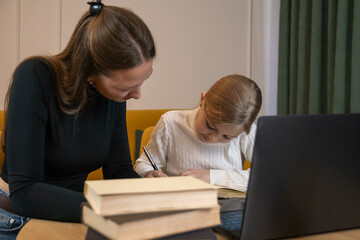 Mother and daughter sitting at a table with a laptop and books, with the parent helping the child study and learn online, fostering academic support and education