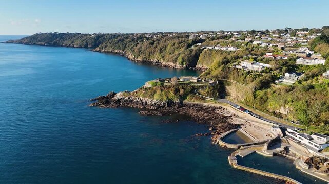 High drone footage pulling back from the seawater bathing pools of Havelet Bay with views over Fort George and eastern coastline of Guernsey on calm day in late afternoon sun