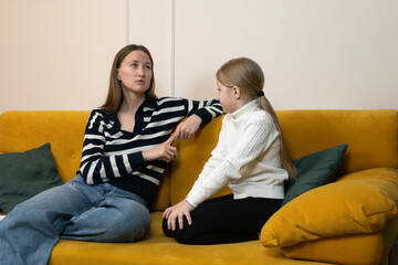 Mother and daughter sit facing each other on a yellow couch at home, sharing a heartfelt conversation, listening and offering support in a candid, emotional family moment
