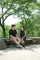 Fototapeta premium A man and woman sit on a bench in a park. The man is drinking from a bottle while the woman is looking at him. Scene is relaxed and intimate