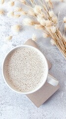 Overhead shot of a cup of coffee with a frothy surface, resting on a coaster, alongside dried flowers, all set against a textured, light-colored background.