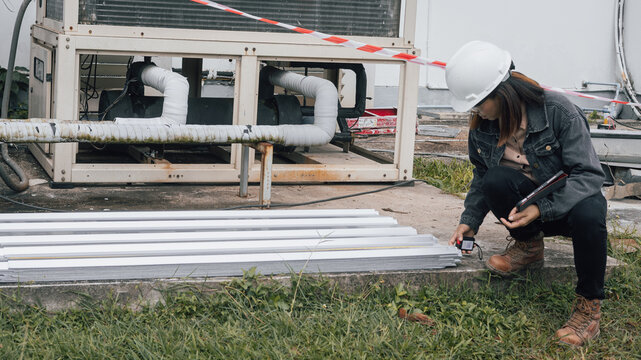 A construction engineer carefully examines metal materials on the ground next to a cooling unit in an outdoor work area. Safety measures are visible in the background - Powered by Adobe