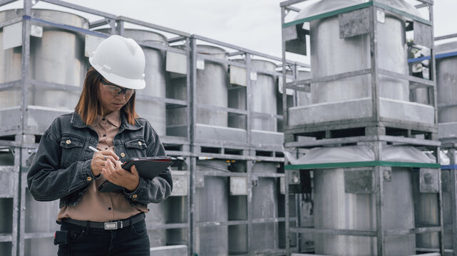 A worker in a hard hat and glasses is taking notes on a tablet while standing among large metal containers at an industrial location. It is daytime, and the sky is cloudy