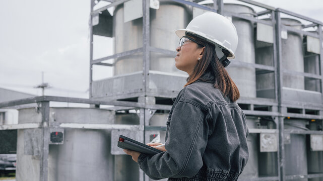 A worker wearing a helmet and glasses stands at a construction site examining machinery. She holds a tablet to track operations and ensure safety during the project
