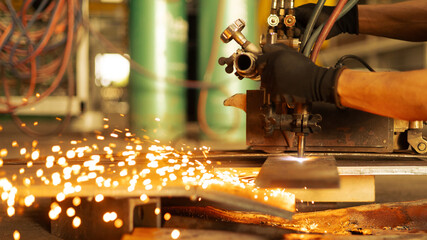A worker is cutting metal with a tool in a workshop. Sparks fly in different directions as the worker focuses on the task. Sunlight streams through the workshop, illuminating the scene