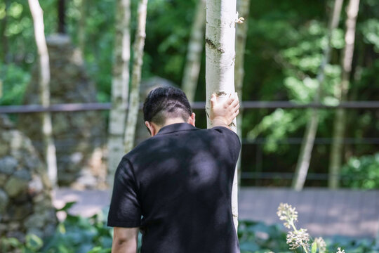 A man is leaning on a tree in a park. The man is wearing a black shirt and he is in a contemplative mood