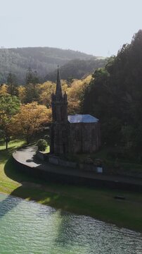 Aerial vertical footage capturing the historic Nossa Senhora das Vit&oacute;rias Chapel on Furnas Lake, S&atilde;o Miguel, Azores. This iconic neo-Gothic landmark is surrounded by lush natural park scener