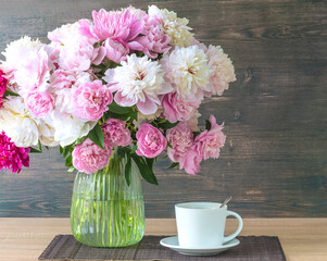 A large bouquet of pink and white peony flowers in glass vase and white cup of coffee or tea on a saucer on a wooden table against dark wooden background
