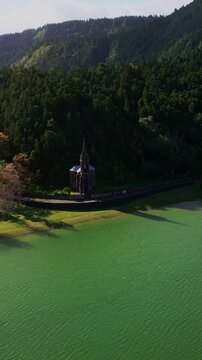 Aerial vertical footage capturing the historic Nossa Senhora das Vit&oacute;rias Chapel on Furnas Lake, S&atilde;o Miguel, Azores. This iconic neo-Gothic landmark is surrounded by lush natural park scener