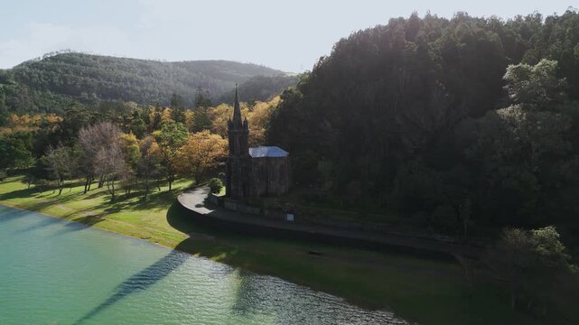 Aerial horizontal footage capturing the historic Nossa Senhora das Vit&oacute;rias Chapel on  Furnas Lake, S&atilde;o Miguel, Azores. This iconic neo-Gothic landmark is surrounded by lush natural park scener