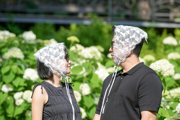 A man and woman are standing in a field of white flowers. The woman is wearing a white headband and the man is wearing a black shirt. They are both smiling and seem to be enjoying each other's company