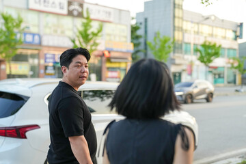 A man and a woman are walking down a street. The man is wearing a black shirt and the woman is wearing a black vest. The street is busy with cars and a truck