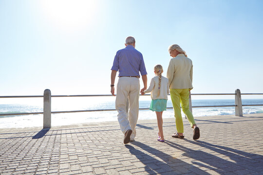 Holding hands, back and grandparents with kid by beach on boardwalk on summer vacation. Travel, walking and senior people with girl child on seaside holiday, weekend trip or getaway together.