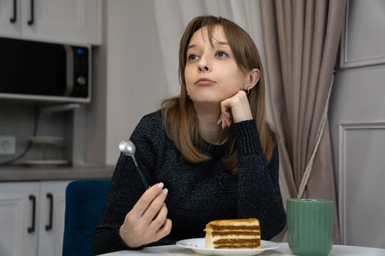 Young woman sits thoughtfully at her kitchen table, holding a spoon and gazing into the distance while a slice of layered cake and a mug rest on the table before her