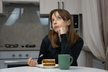 Young woman sitting thoughtfully at kitchen table holding spoon and looking up while piece of layer cake and mug lying on table in front of her