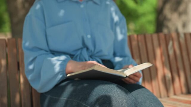 smiling caucasian student reading under tree on wooden bench, candid portrait with dappled light, denim skirt and soft blouse, attentive expression turning pages, calm campus vibe and gentle warmth