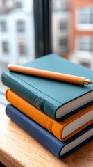 Close-up shot of a stack of three colorful notebooks with a pen resting on top, placed on a wooden surface near a window. The background shows blurred buildings