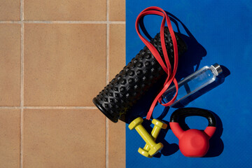 Set of fitness equipment on yoga mat including dumbbells, kettlebell, resistance band, foam roller, and water bottle outdoors