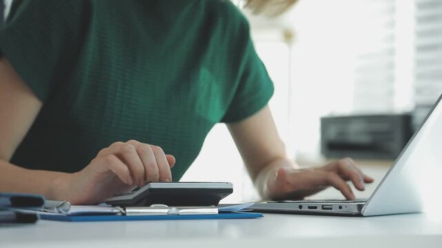 Two financial accountants analysing business spreadsheets on laptop computer, sitting at office desk, looking through paper documents, using calculator working with databases, checking excel tables