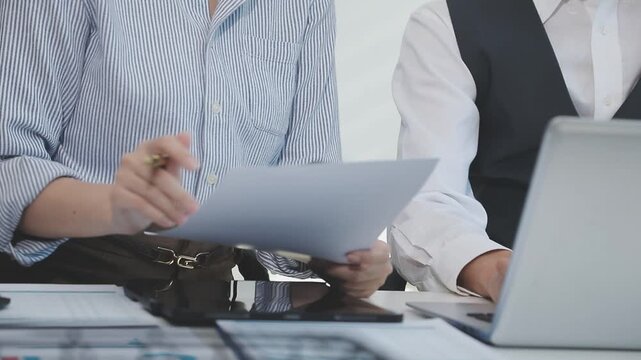 Two financial accountants analysing business spreadsheets on laptop computer, sitting at office desk, looking through paper documents, using calculator working with databases, checking excel tables