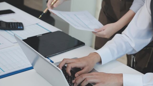 Two financial accountants analysing business spreadsheets on laptop computer, sitting at office desk, looking through paper documents, using calculator working with databases, checking excel tables