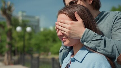 man covers eyes of white woman while boyfriend surprises girlfriend with playful kiss and warm embrace in sunny urban park, denim jacket, tree-lined walkway, intimate candid moment full of laughter