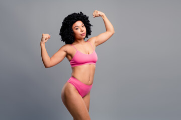 Confident young woman in pink bikini flexes arms showing strength and fitness against a grey...