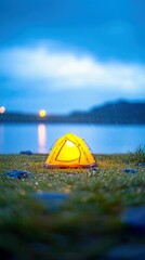 A yellow tent glows from within, set on a grassy bank near a lake, with rain falling under a dusky sky.