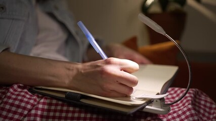 Close-up of a woman writing her thoughts, plans, or memories in a personal diary at night, illuminated by a small portable reading lamp clipped to the page for focused light