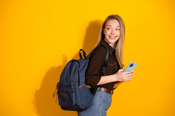 Young woman with backpack smiling while using her phone against a vibrant yellow background