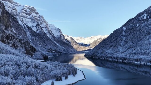 Scenic aerial view of Kl&ouml;ntalersee, Glarus, Switzerland. Snow-dusted mountains meet the dark, calm lake, creating a perfect, striking reflection of the Kl&ouml;ntal valley under clear skies.