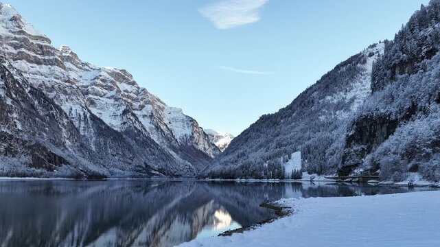 A serene winter look at Kl&ouml;ntalersee in Glarus, Switzerland, where a dark, motionless lake mirrors the steep, snow-draped Kl&ouml;ntal mountains beneath a crisp, clear blue sky.