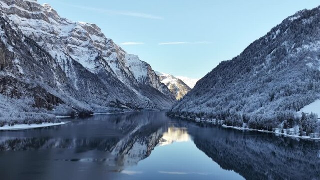 A pristine winter scene at Kl&ouml;ntalersee in Glarus, Switzerland, where the snowy shoreline meets a dark, glass-still lake reflecting the towering Kl&ouml;ntal valley beneath a bright, clear sky.