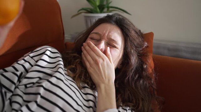 Young pregnant woman feeling nauseous, smelling a lemon to alleviate the symptoms of morning sickness during her first trimester while resting on a couch, clearly showing her distress