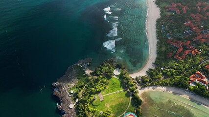 Aerial drone footage of the peninsula at Bali during golden hour showing turquoise lagoon beside a sandy beach with a coastal park and resort area while open ocean surf breaks along the shoreline - Powered by Adobe