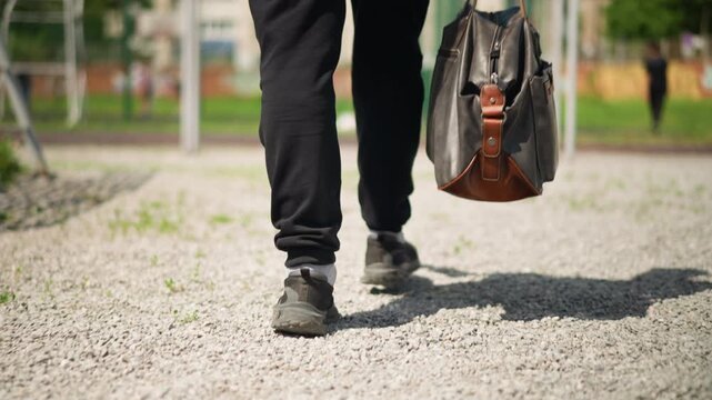 city dweller with leather bag, person walking on gravel with urban surroundings behind, male individual confidently strolling along gravel path with city backdrop and leather accessories