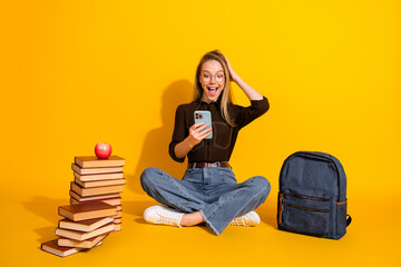 Young woman student phone smiling sits on floor stack of book backpack yellow background for...