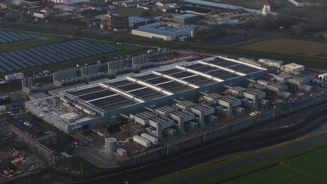 Aerial drone video, massive industrial-scale data center building exterior in Winschoten, The Netherlands, roof covered with solar panels generating renewable energy for computing and AI hardware.
