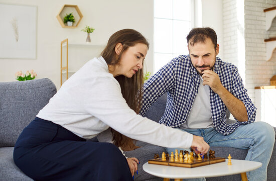 Couple playing chess together at home, people planning strategy and tactics to win competition. Husband and wife play smart game, sitting on sofa by chessboard to think and focus on next move