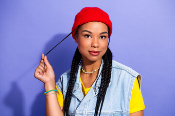 Young woman with red beanie and braids in denim vest and yellow shirt posing in urban style against purple background for fashion lifestyle streetwear and advertising