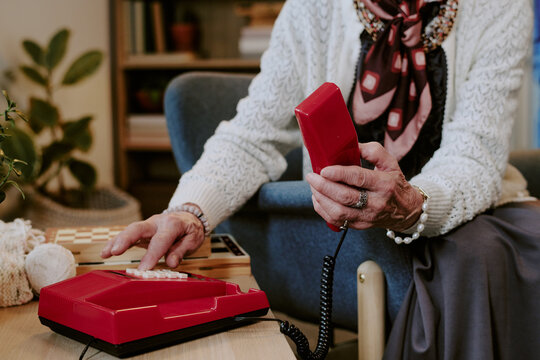 Senior Caucasian woman sitting in living room holding red landline telephone receiver in one hand while dialing number with other hand, wearing patterned scarf and knit sweater