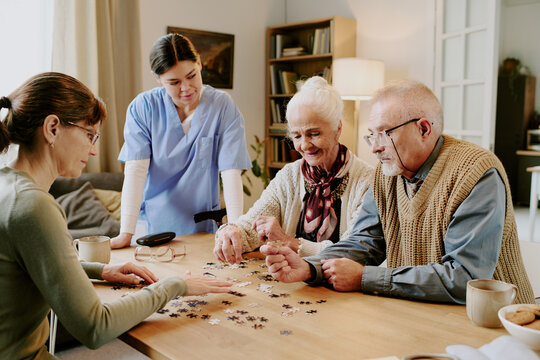 Senior Caucasian woman and senior man assembling puzzle pieces at table with middle aged woman and young adult female caregiver observing interaction