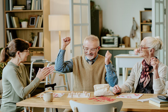 Senior Caucasian man celebrating chess victory with arms raised, while senior Caucasian woman and middle aged Caucasian woman sitting at table applauding and smiling together