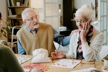 Senior Caucasian man comforting senior Caucasian woman sitting at table playing bingo, woman holding hand to face appearing emotional, man placing hand on her shoulder, game pieces scattered