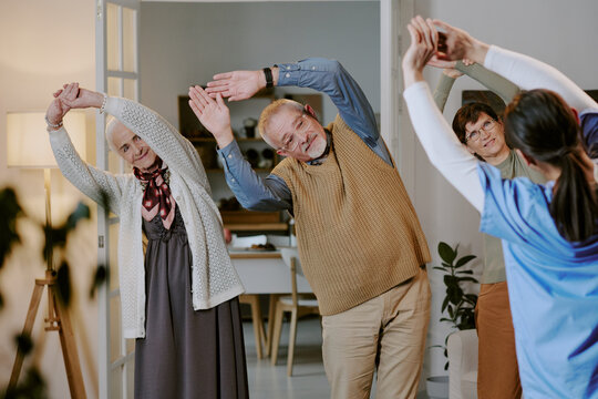 Group of senior Caucasian man and women following young adult female instructor stretching arms during fitness class indoors, all participants standing and engaging in physical exercise together - Powered by Adobe