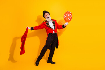 Circus showman in red coat and top hat grins with balloon on bright yellow background celebrating performance and joy