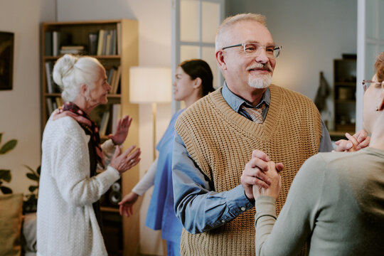 Senior Caucasian man smiling while dancing with senior Caucasian woman in living room, two senior women talking and gesturing in background, group enjoying social interaction indoors
