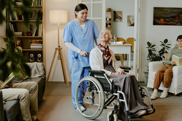 Senior Caucasian woman with disability sitting in wheelchair smiling at young adult Hispanic female...